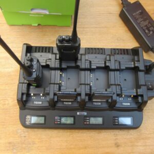 The Quansheng TG330 Twelve Slot Charger, with two walkie-talkies docked and three empty slots, sits on a wooden surface beside a power adaptor.
