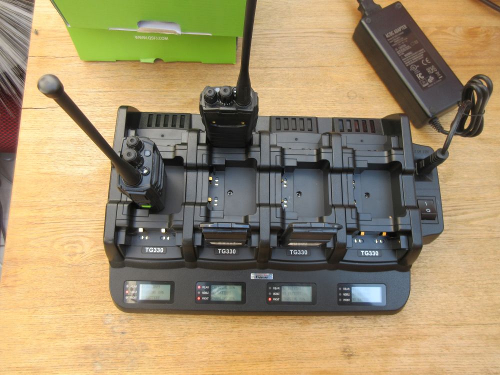 The Quansheng TG330 Twelve Slot Charger, with two walkie-talkies docked and three empty slots, sits on a wooden surface beside a power adaptor.