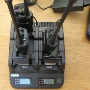 Two TG330 walkie-talkies sit in a Quansheng TG330 Six Slot Charger with digital displays, resting on a wooden surface.