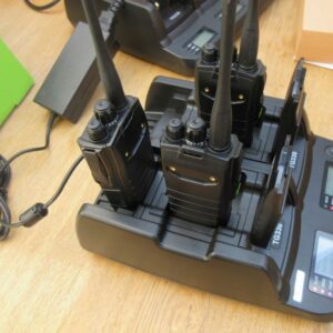 Four handheld two-way radios stand upright in a Quansheng TG330 Six Slot Charger on a wooden table, with their digital screens showing charging status.