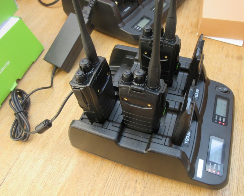 Four handheld two-way radios stand upright in a Quansheng TG330 Six Slot Charger on a wooden table, with their digital screens showing charging status.