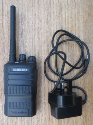 amherst-a66-walkie-talkie-with-plugin-charger-m An Amherst A66 Compact Walkie-Talkie and its black power adaptor with lead are placed side by side on a wooden surface.