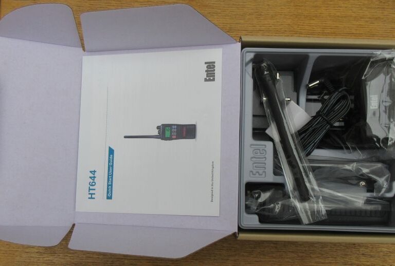 Open box containing an Entel HT644 Marine VHF Walkie Talkie set with instruction manual, handheld radio, aerial, charging base, and power adaptor on a wooden surface.
