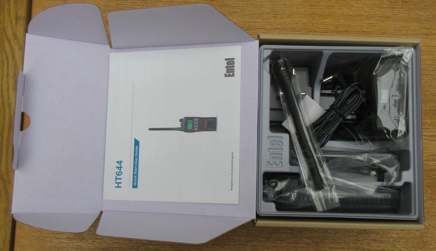 entel-ht644-box-contents Open box containing an Entel HT644 Marine VHF Walkie Talkie set with instruction manual, handheld radio, aerial, charging base, and power adaptor on a wooden surface.