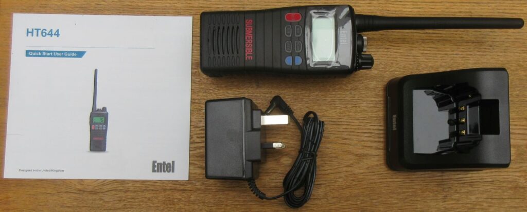 entel-ht644-what-you-get The Entel HT644 Marine VHF Walkie Talkie set, including a handheld radio, quick start guide, power adaptor, and charging dock, is displayed on a wooden surface.