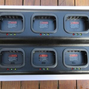 The Kirisun PT568 Six Slot Multi-Charger, featuring indicator lights and warning labels on each slot, sits on a wooden surface. Each slot allows for easy battery insertion.