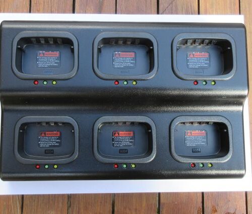 kirisun-six-slot-charger-on-a4-paper (1) The Kirisun PT568 Six Slot Multi-Charger, featuring indicator lights and warning labels on each slot, sits on a wooden surface. Each slot allows for easy battery insertion.