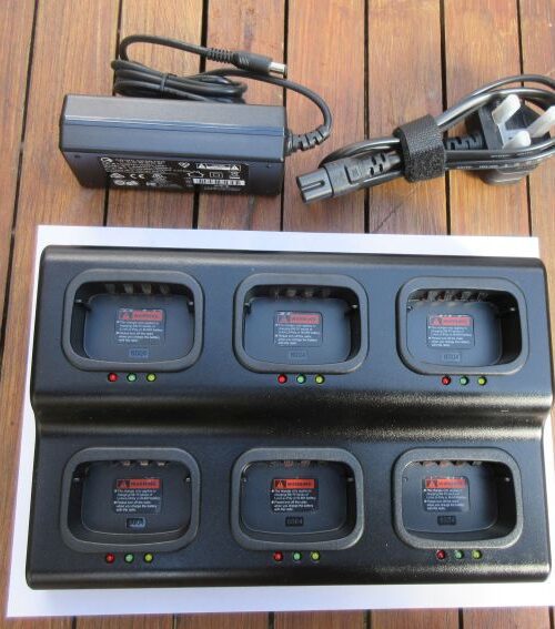The Kirisun PT558 / PT4200 Six Slot Multi-Charger, supplied with a mains adaptor and power lead, rests on a wooden surface. Each of its six slots features indicator lights and warning labels.