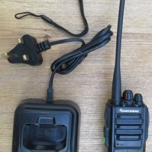 quansheng-tg330-walkie-talkie-with-charger-m A Quansheng TG330 Walkie Talkie Radio with a long aerial, charging dock, and mains plug are displayed on a wooden surface.