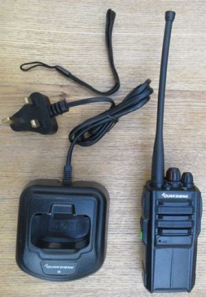 quansheng-tg330-walkie-talkie-with-charger-m A Quansheng TG330 Walkie Talkie Radio with a long aerial, charging dock, and mains plug are displayed on a wooden surface.