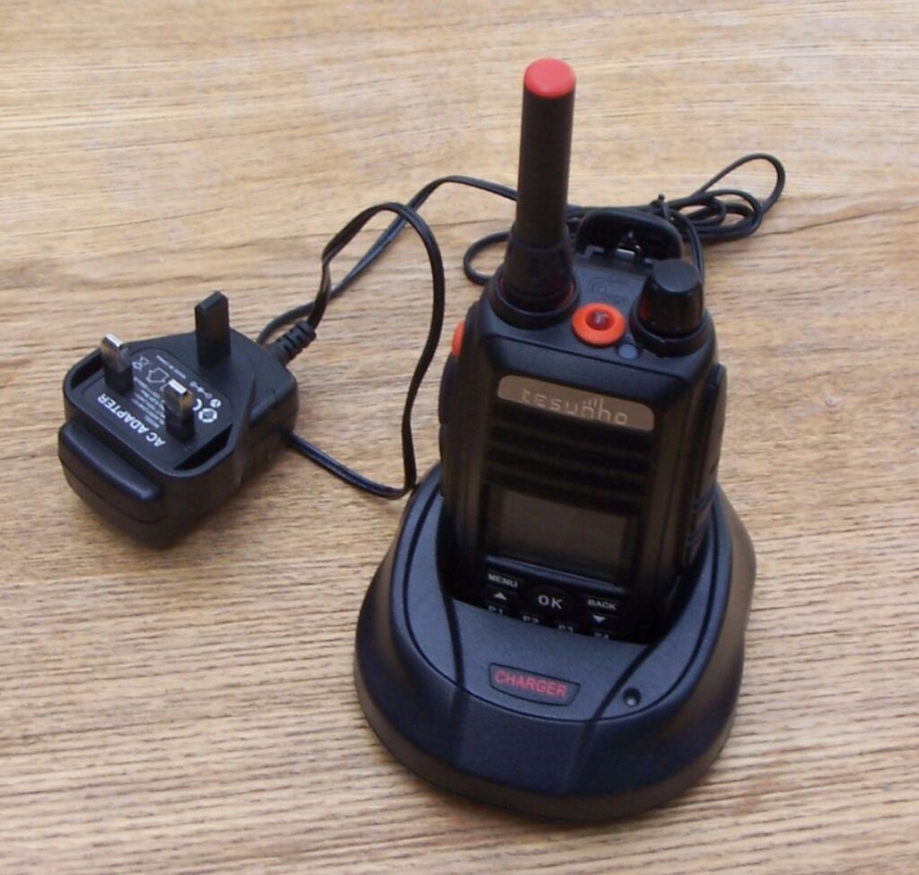A Tesunho TH388 Display GPS Network Walkie-Talkie Radio sits in its charging dock on a wooden surface, with the power adaptor plugged in beside it.