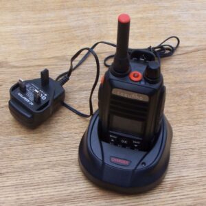 A Tesunho TH388 Display GPS Network Walkie-Talkie Radio sits in its charging dock on a wooden surface, with the power adaptor plugged in beside it.