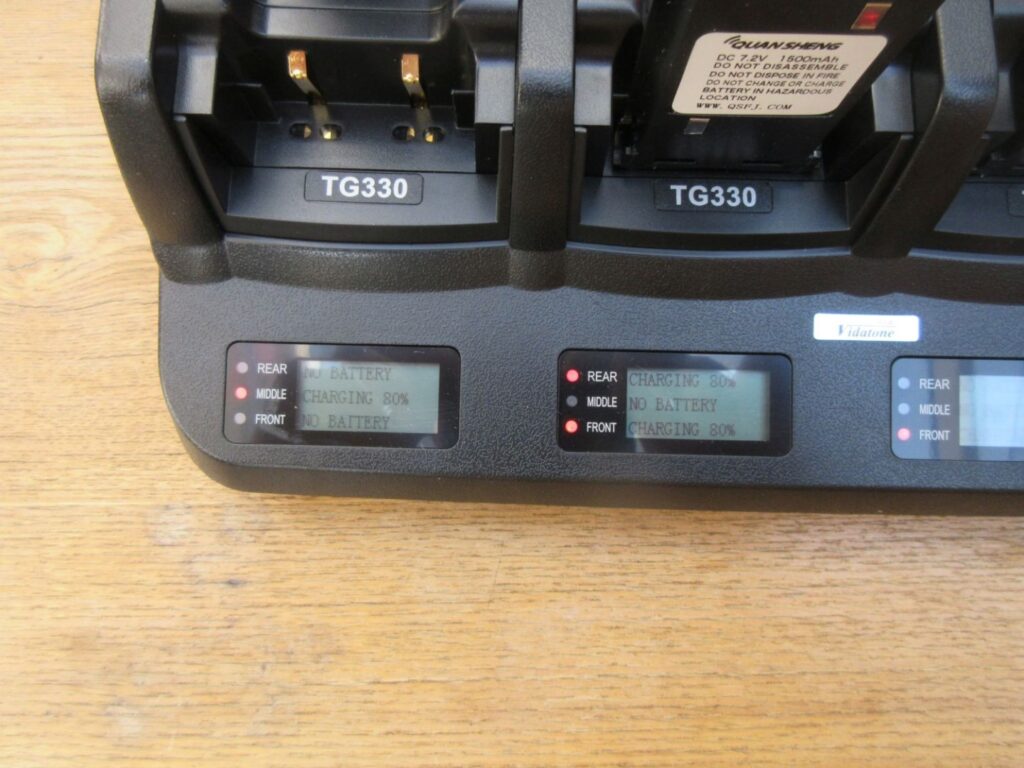 A close-up of the Quansheng TG330 Six Slot Charger with digital screens displaying "CHARGING 80%" and "NO BATTERY" status indicators.